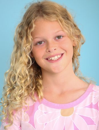 Smiling young girl with long curly blonde hair poses indoors against a blue backdrop wearing a pink shirt with white daisy flowers.