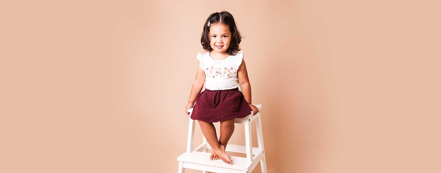 A happy young girl with curly hair and a hair clip is sitting on a white stool in front of a peach-colored background in a studio shot.