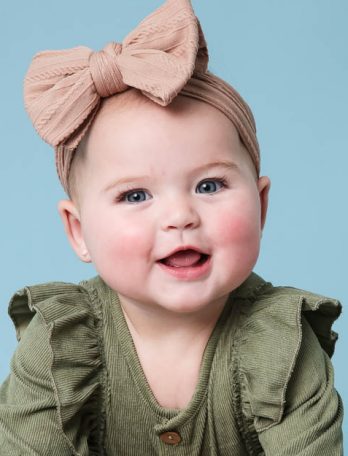 Portrait of a baby with a large bow in her hair, smiling, wearing a green outfit, on a light blue background. Close up shot.