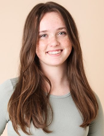 Portrait of a young woman with long brown hair and freckles smiling in a studio setting with a plain beige background. Close-up shot.