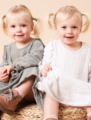 Two young girls with blonde hair in pigtails, wearing dresses and shoes, sitting close together on a wicker surface indoors.