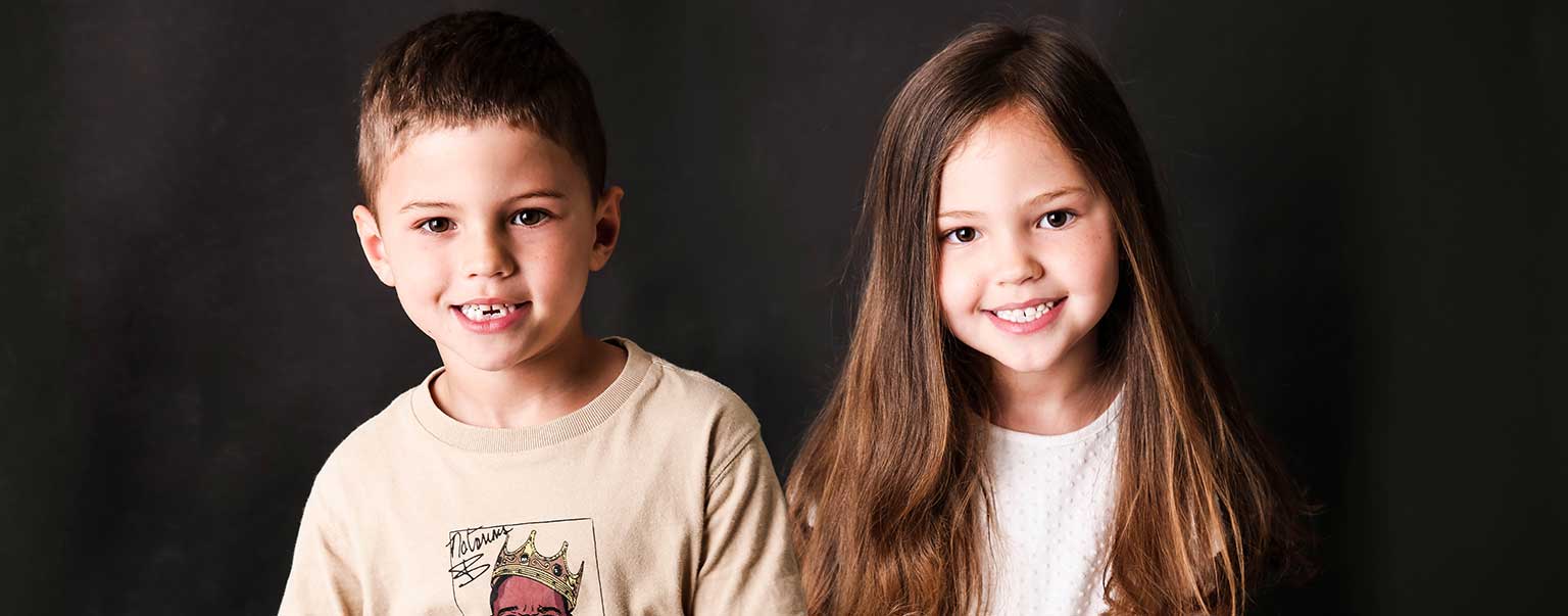 Portrait of smiling young boy and girl side-by-side against a dark backdrop. They are wearing casual tops, looking directly at the camera.
