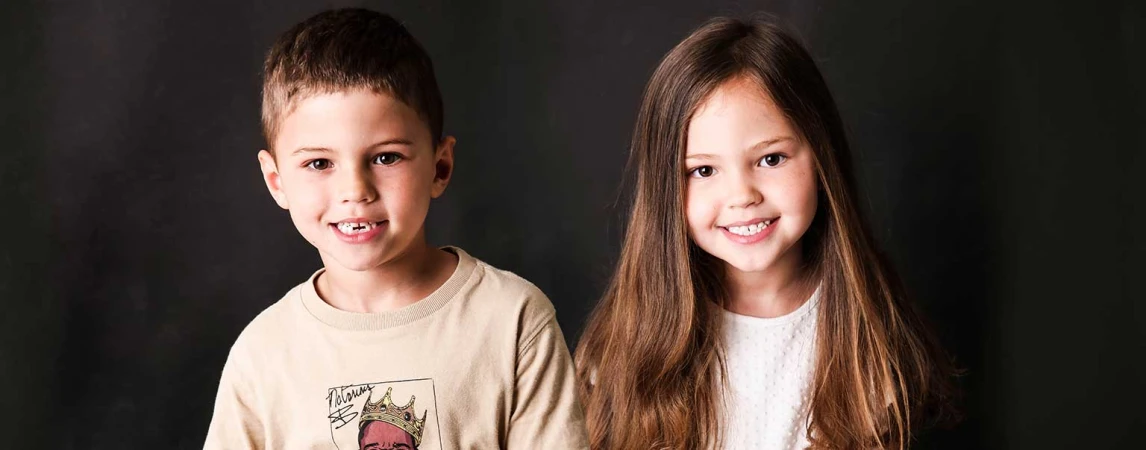 Portrait of smiling young boy and girl side-by-side against a dark backdrop. They are wearing casual tops, looking directly at the camera.