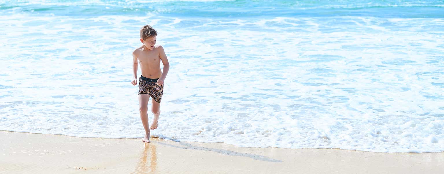 Boy runs in patterned swim trunks at ocean's edge on bright, sunny day with blue water and waves, golden sand, and clear horizon.
