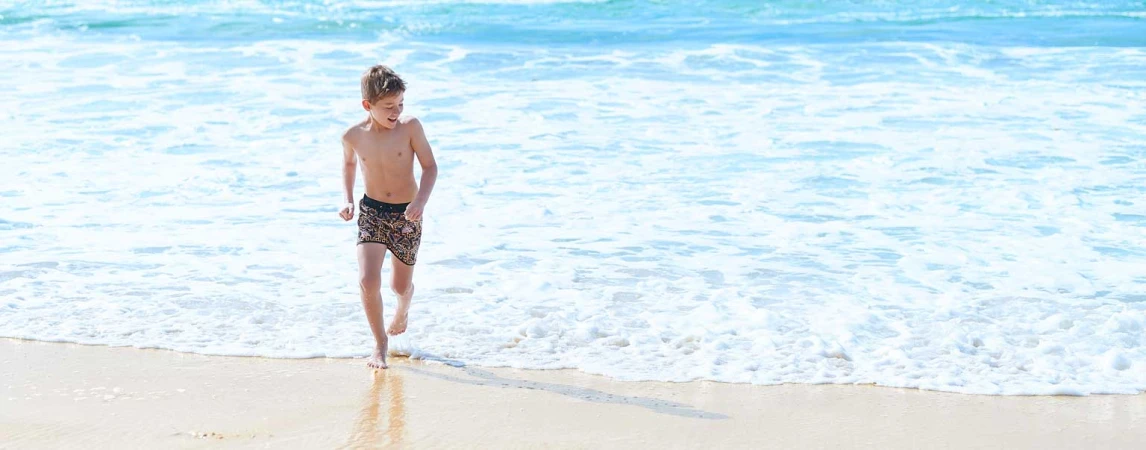 Boy runs in patterned swim trunks at ocean's edge on bright, sunny day with blue water and waves, golden sand, and clear horizon.