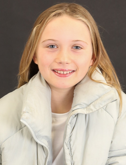 Portrait of a smiling young girl with long blonde hair, wearing a light-blue puffer jacket against a dark-grey studio backdrop.