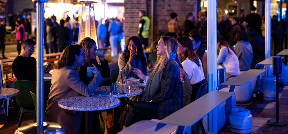 Outdoor cafe scene at night features a group of women at tables, illuminated by soft lighting and patio heaters, with a bustling crowd.