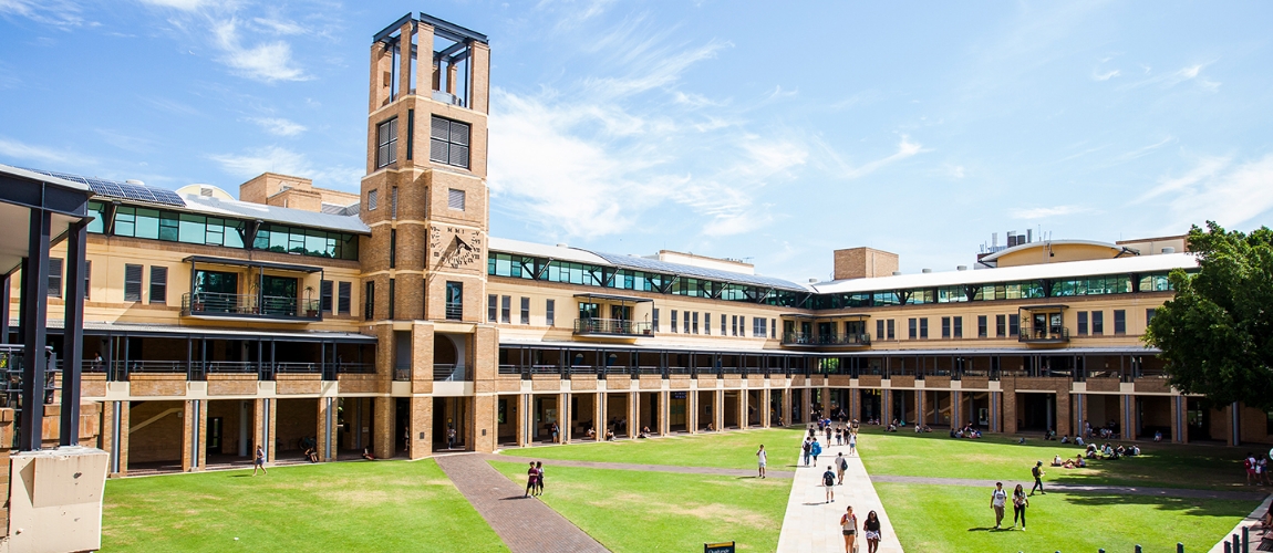 Exterior view of a multi-story tan building with a clock tower on a college campus, with many people walking along the lawn.