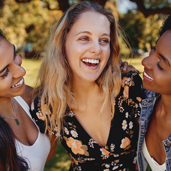 Three smiling young women embrace outdoors on a sunny day. One woman is blonde, one has dark hair, and one is light-skinned.
