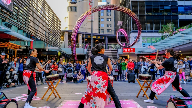 Three Japanese drummers perform in a city square, surrounded by a crowd and a large spiral sculpture, showcasing cultural entertainment.
