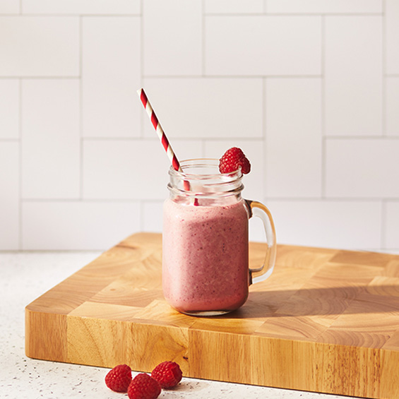 Raspberry smoothie in a mason jar with a red and white striped straw, sitting on a wooden cutting board. White tile background.
