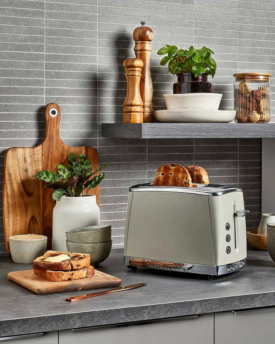 Kitchen still life featuring a beige toaster with bread, a cutting board with toast, bowls, and wooden accents against a gray backsplash.