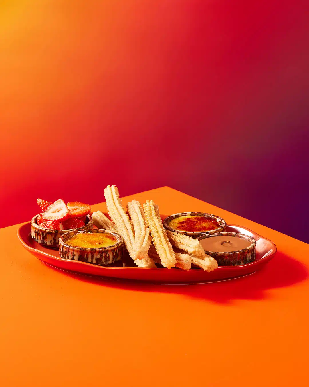 Overhead shot of a vibrant orange table featuring churros, various dipping sauces in ramekins, and fresh strawberries on a red plate.