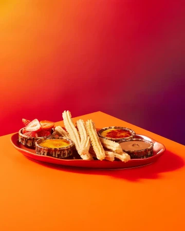 Overhead shot of a vibrant orange table featuring churros, various dipping sauces in ramekins, and fresh strawberries on a red plate.