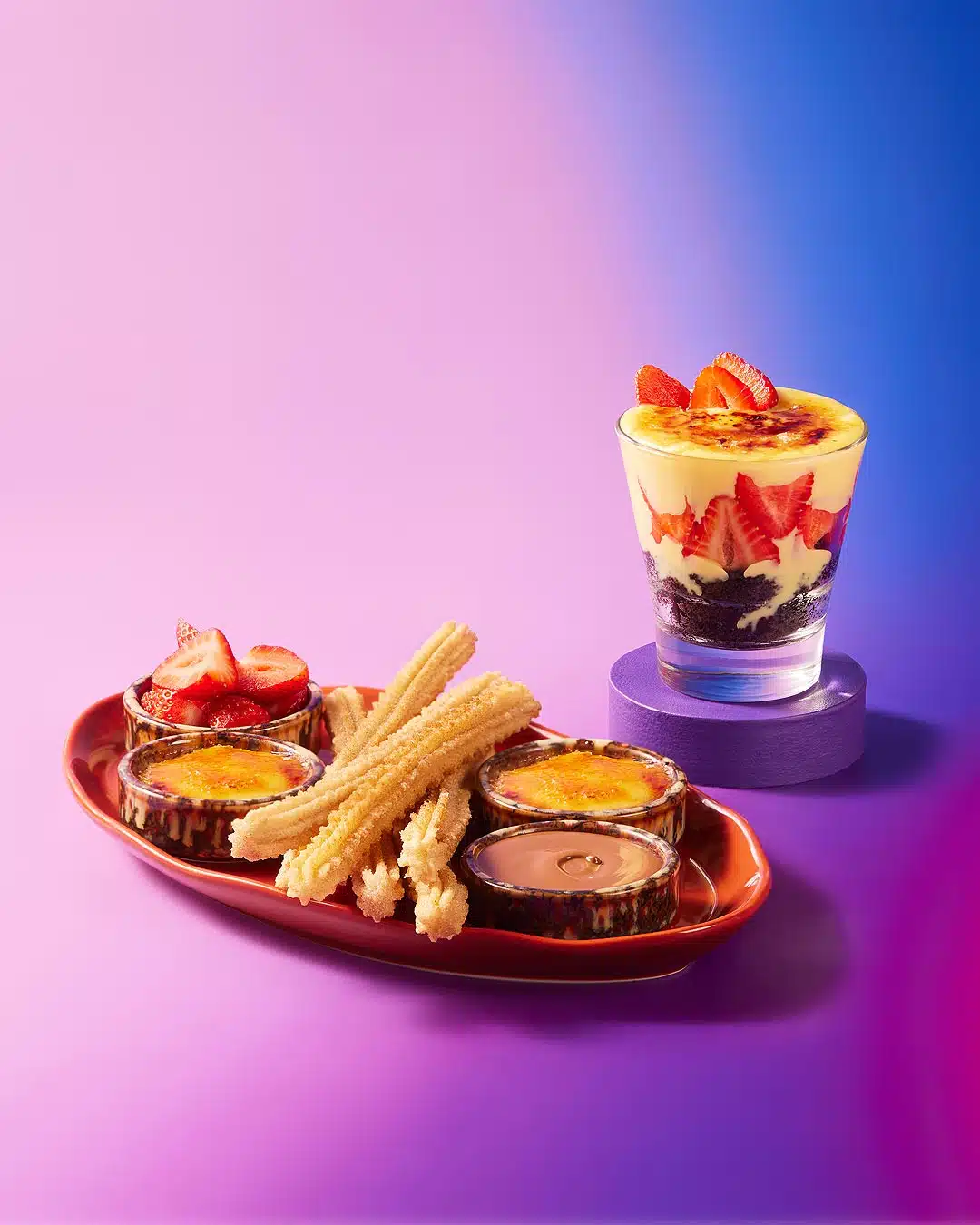 Elegant dessert platter featuring churros, crème brûlée variations, and a layered strawberry parfait, beautifully presented against a dual-toned backdrop.