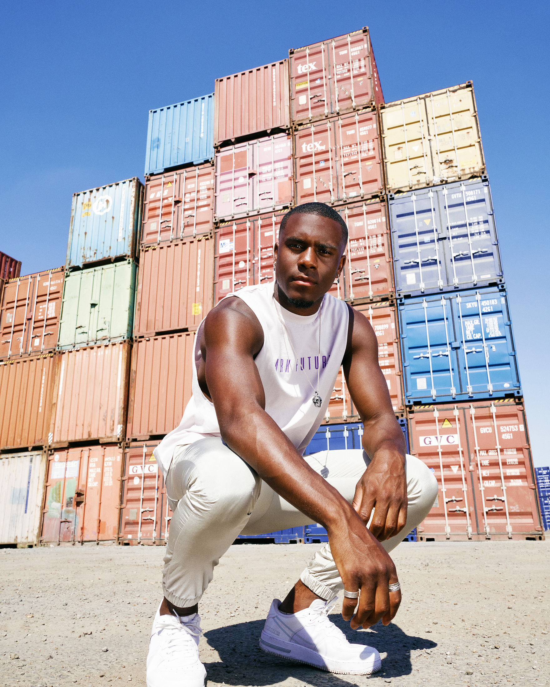 A fit dark-skinned man in a white tank top and matching pants crouches in front of a stack of colorful shipping containers.