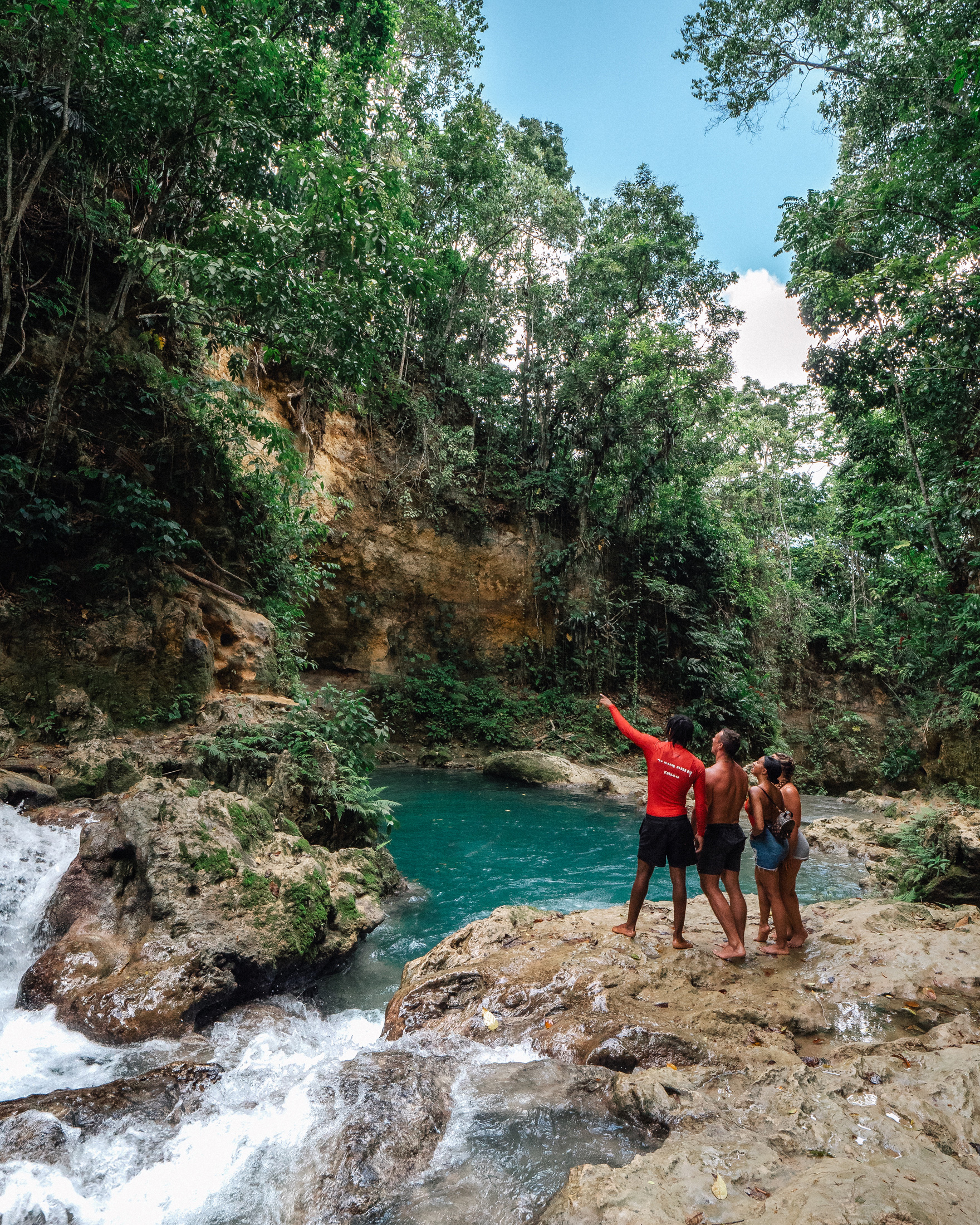 A group of people stands on rocks overlooking a turquoise pool and waterfall surrounded by lush green trees under a bright blue sky.