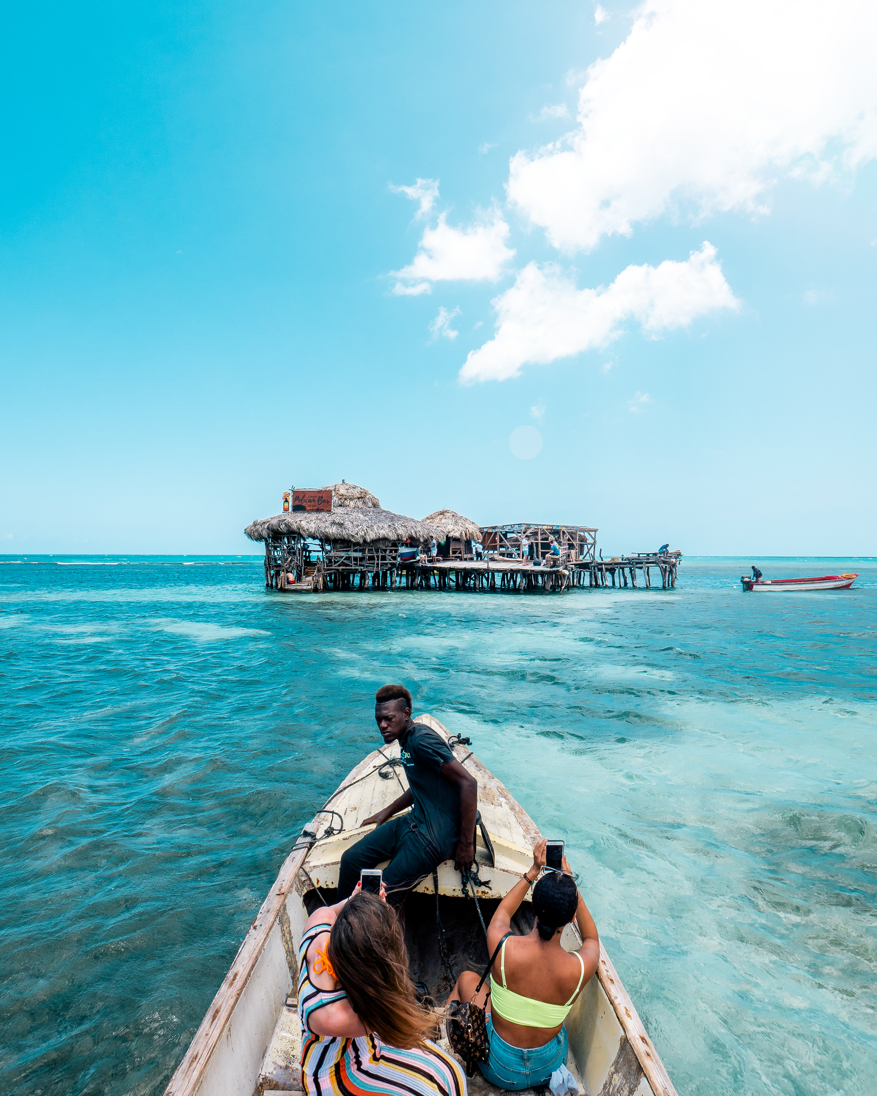 Group of people riding in a boat towards a thatched-roof structure on the water under a blue sky with scattered clouds, tropical destination.