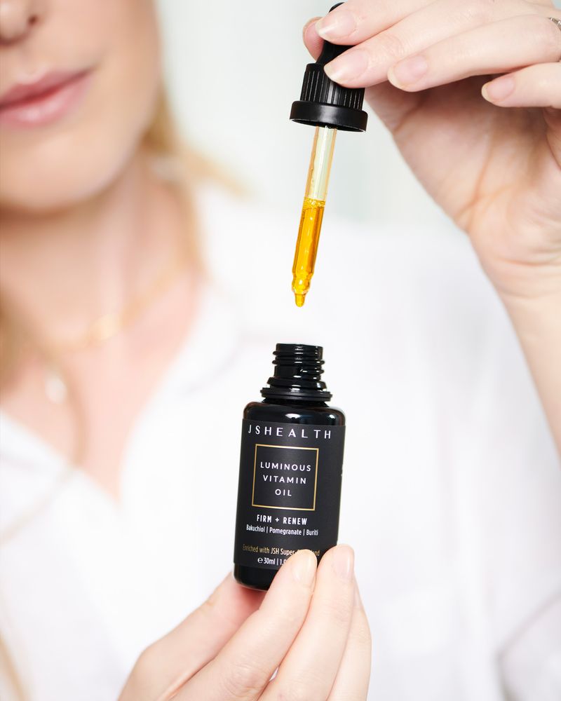Close-up of a woman's hands holding a bottle of luminous vitamin oil and a dropper with golden liquid in an indoor setting.