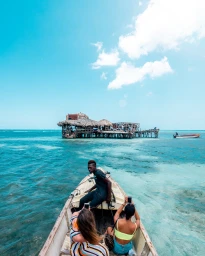 Three people riding in a small wooden boat on bright turquoise water with a stilt hut in the distance under a blue sky.