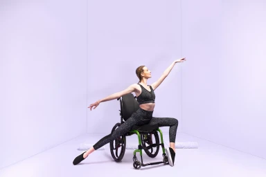 Dancer in black athletic wear seated in a green wheelchair striking a graceful pose in a light purple studio.