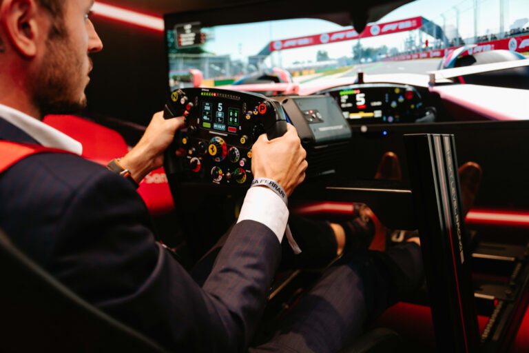 Man wearing a suit holding a steering wheel in a driving simulator with a racetrack visible on a monitor in the background.