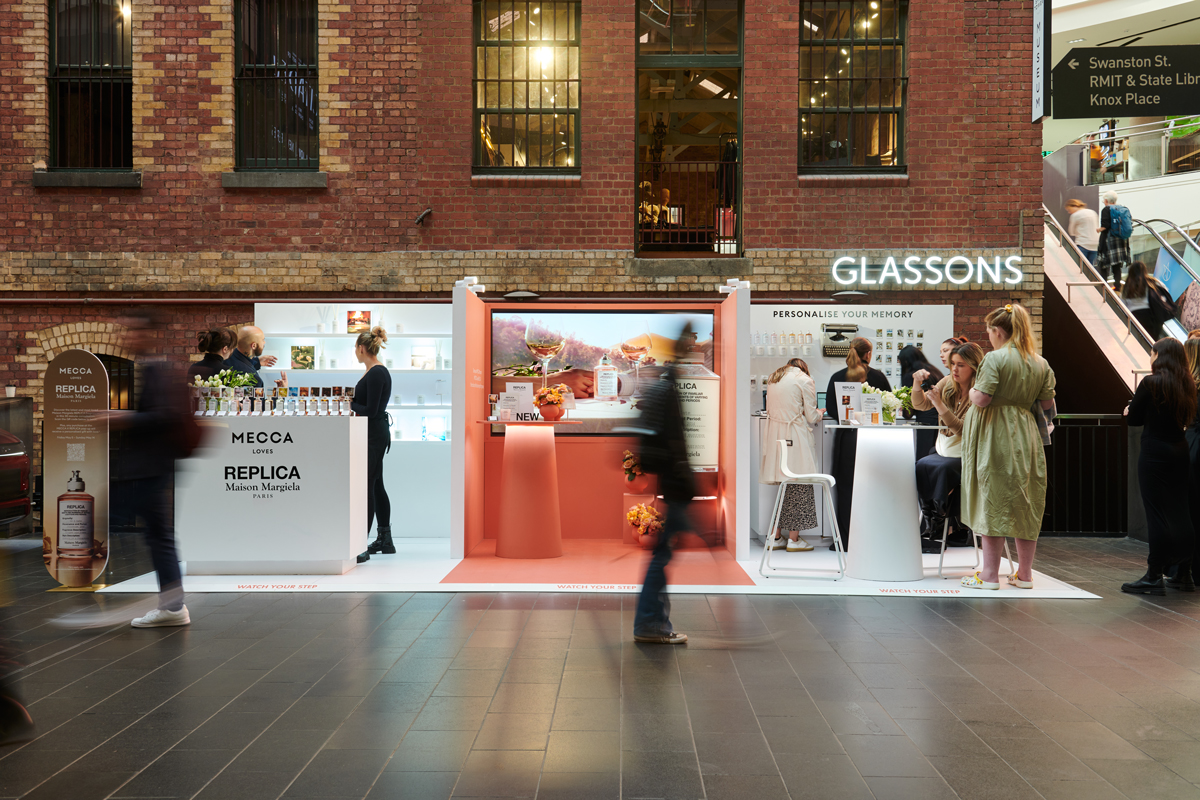 Indoor shopping mall featuring pop-up cosmetics stands, displays with makeup and skincare products, and several people browsing the beauty items.