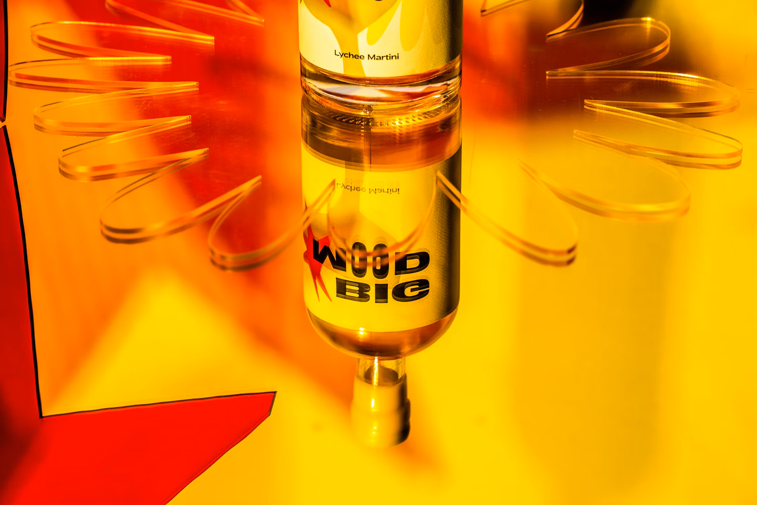 A bottle of Lychee Martini liquor sits on a yellow surface surrounded by transparent, petal-shaped plastic pieces creating a flower effect.