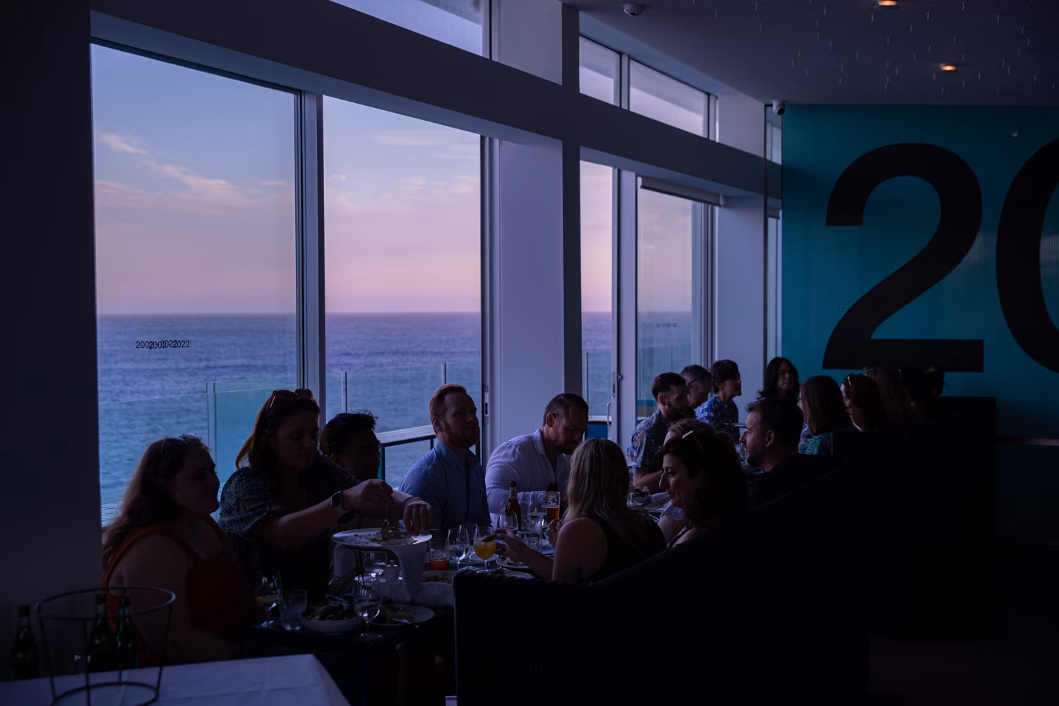 A group of people dining indoors near a window with an ocean view at dusk, creating a dimly lit and intimate gathering.