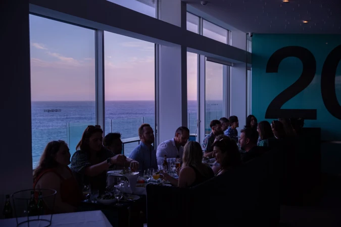 A group of people dining indoors near a window with an ocean view at dusk, creating a dimly lit and intimate gathering.