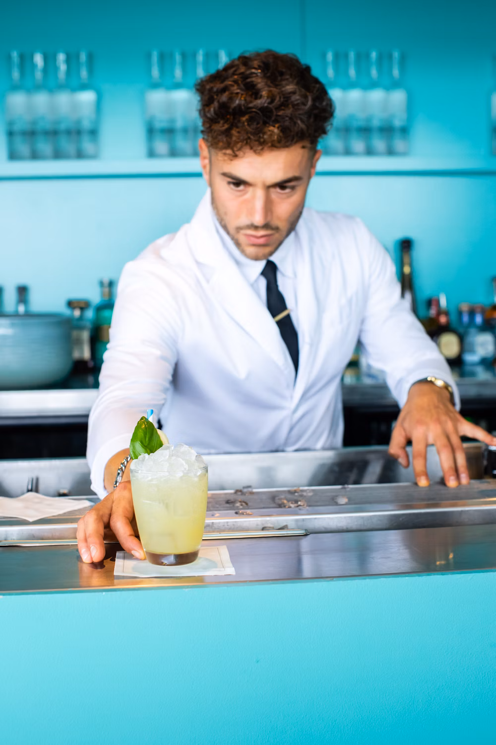 A bartender in a white coat places a cocktail on a metal bar, featuring a light-yellow drink with crushed ice and a green garnish.