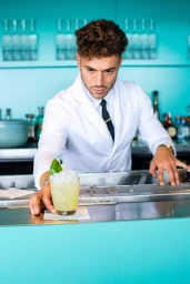 Bartender in a white chef coat and black tie serving a cocktail on a turquoise bar counter
