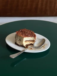 Slice of tiramisu with a silver spoon on a white oval plate resting on a dark green tabletop