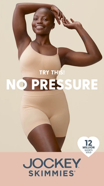 Smiling African-American woman poses in Jockey Skimmies underwear. Studio shot of adult female in shapewear. Tan colored background.