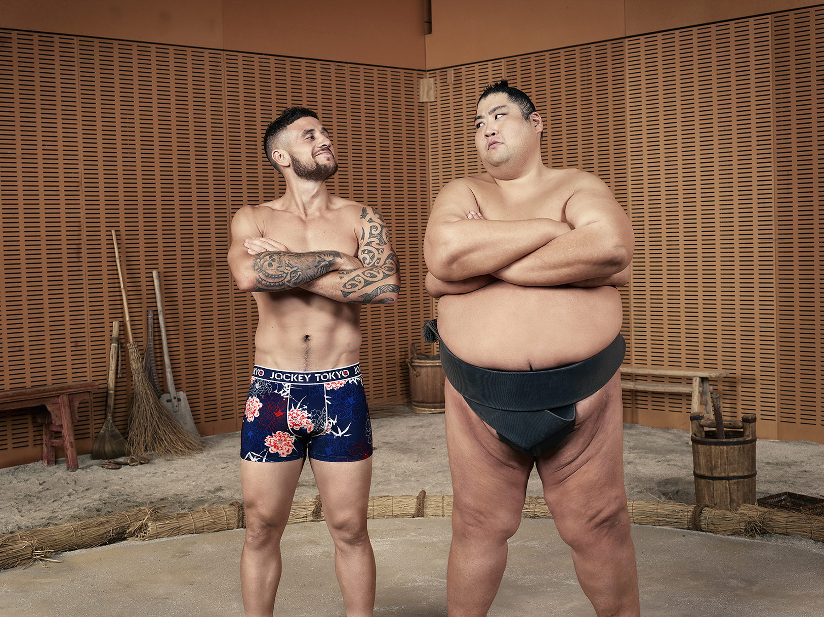 Two men pose in a sumo ring: one muscular and tattooed in floral boxers, the other a sumo wrestler in traditional mawashi.