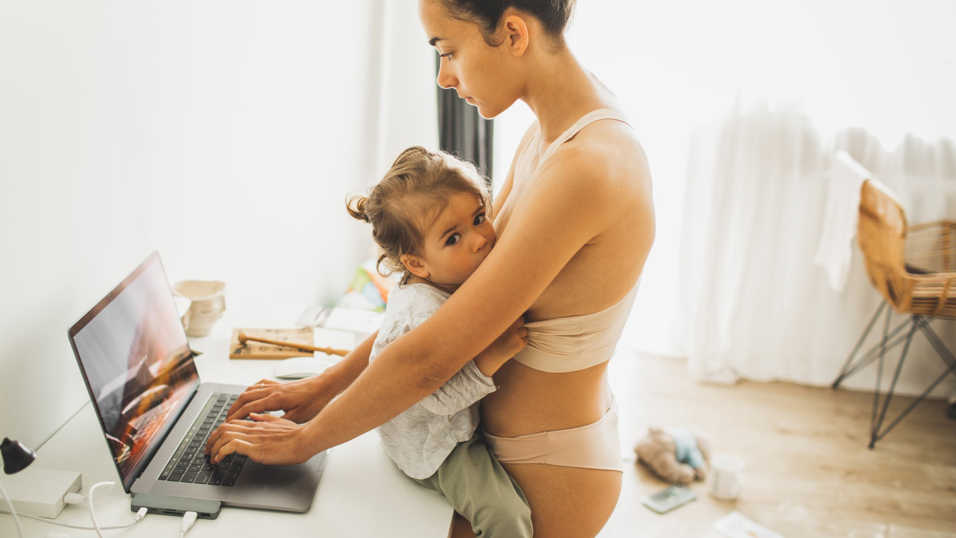 Young mother breastfeeds toddler while working from home on a laptop. The image captures the blend of motherhood and modern work life.