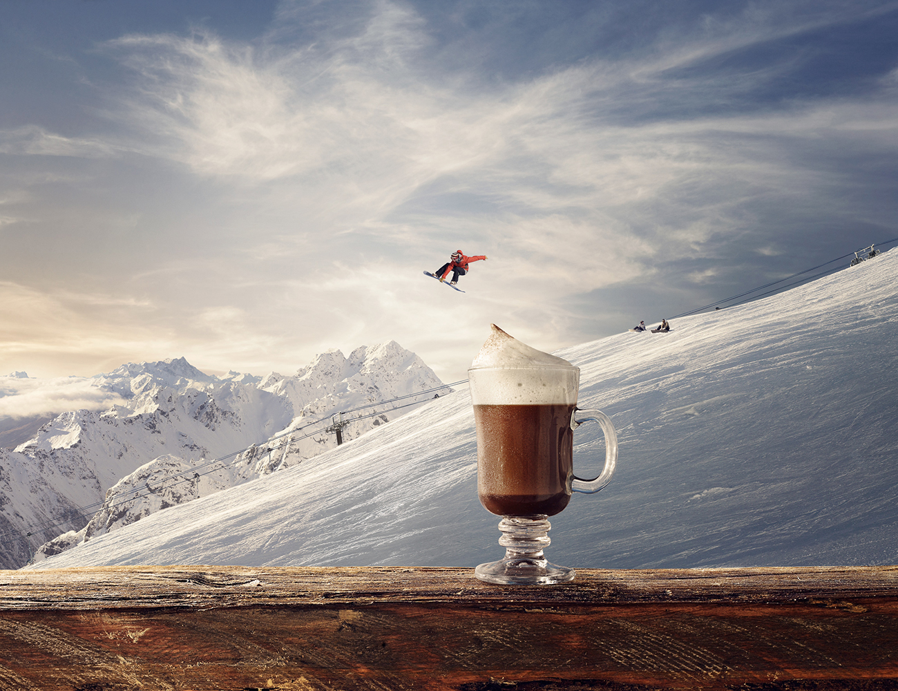 Hot chocolate with cream in a glass on a wooden table, with a snowboarder in the background jumping against a snowy mountain landscape.