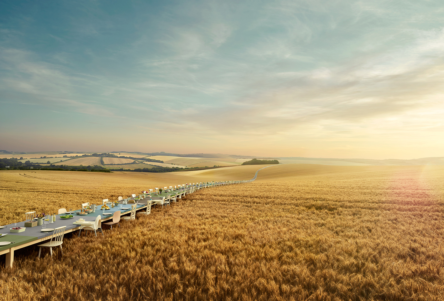 A surreal outdoor dining experience features an extremely long table set in a golden field under a blue sky, creating a dreamlike atmosphere.