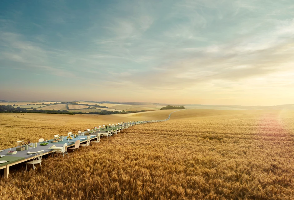 A surreal outdoor dining experience features an extremely long table set in a golden field under a blue sky, creating a dreamlike atmosphere.