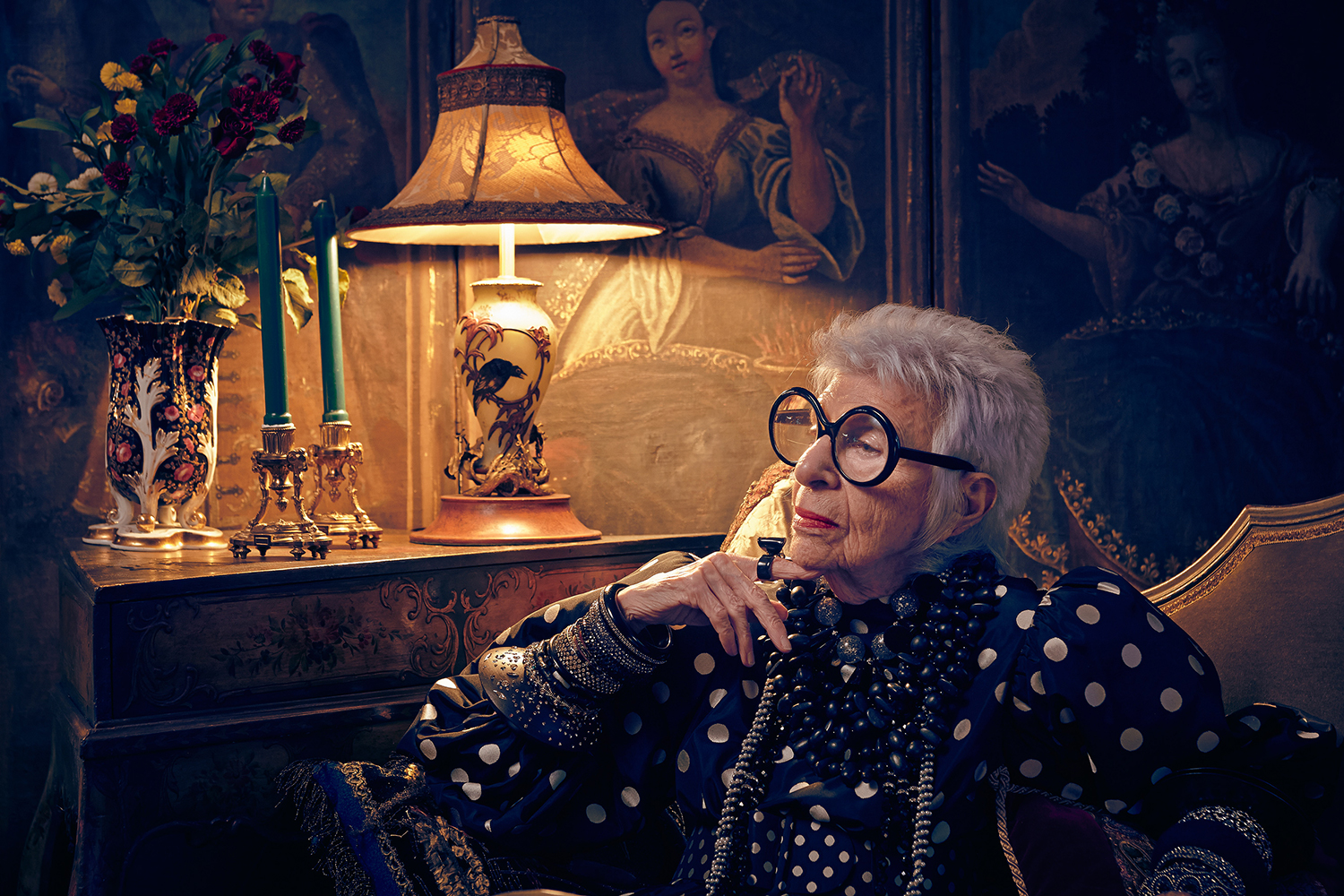 Elegant elderly woman with white hair and large glasses poses in an armchair in a richly decorated room with vintage paintings.