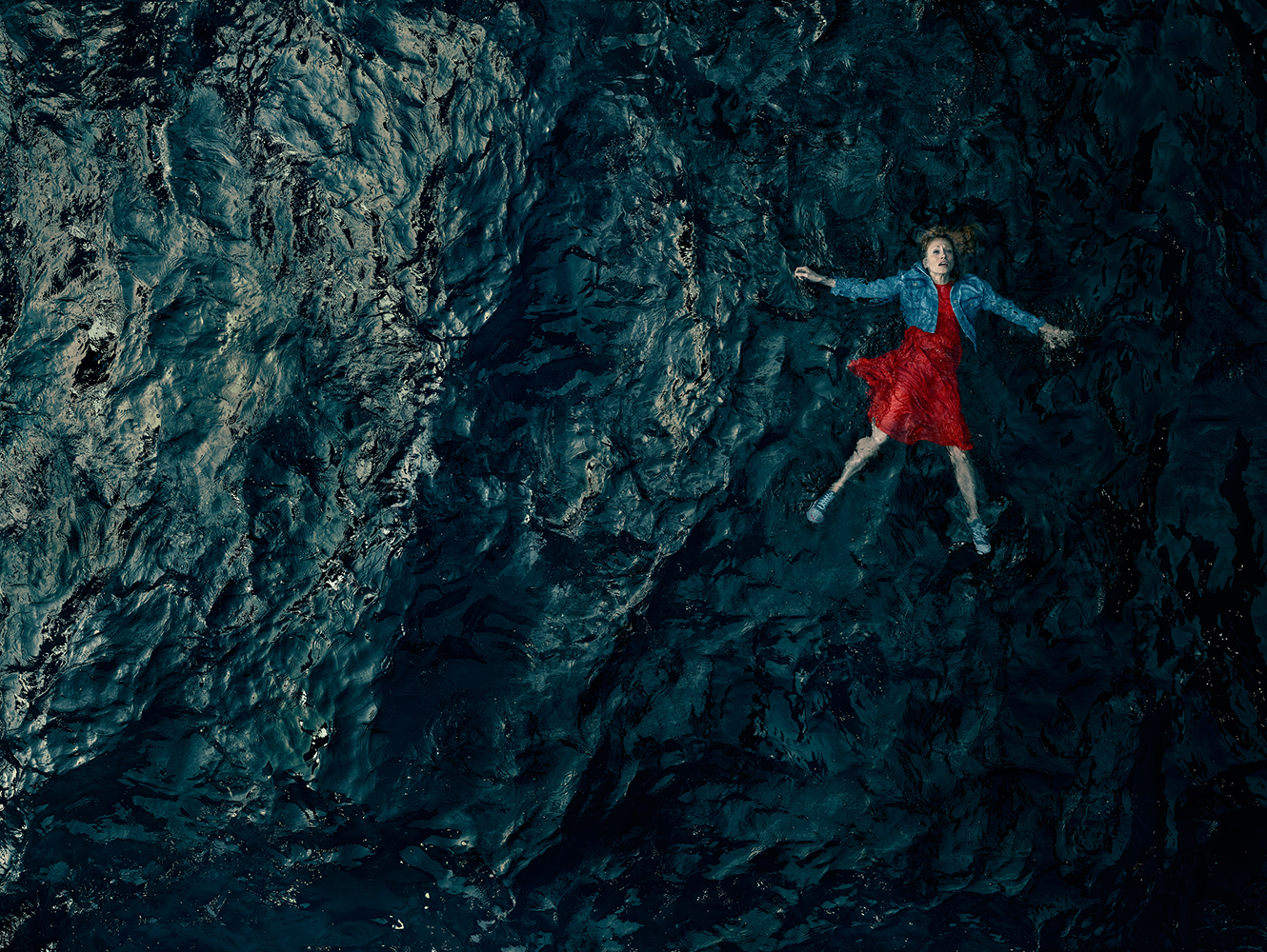 Aerial view of a woman in a red dress and jean jacket floating on her back in dark water with ripples and reflections, creating a serene image.