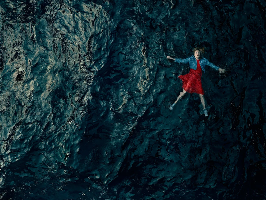Aerial view of a woman in a red dress and jean jacket floating on her back in dark water with ripples and reflections, creating a serene image.