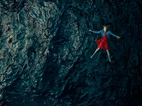 Aerial view of a woman in a red dress and jean jacket floating on her back in dark water with ripples and reflections, creating a serene image.