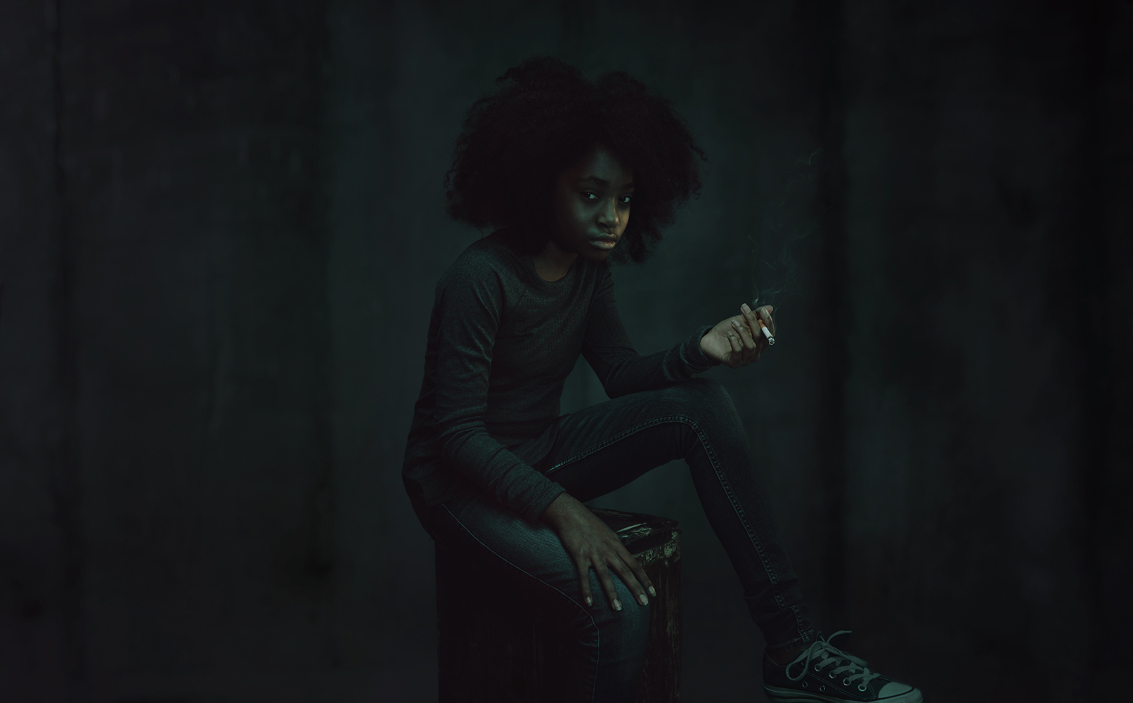 An African American woman sits in a dimly lit room, wearing a long-sleeved top and jeans, while holding a lit cigarette.