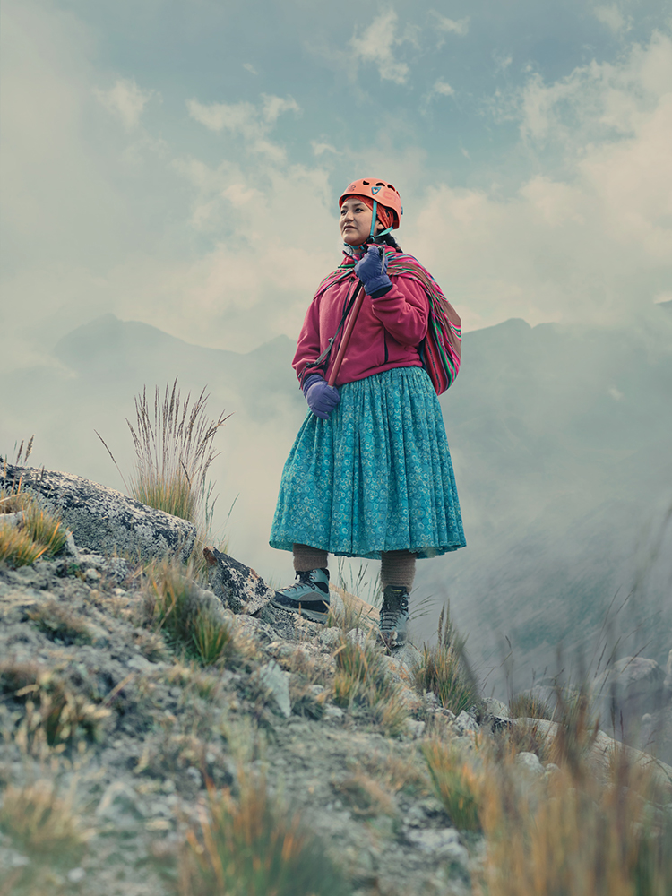 Adult female mountain climber stands on a rocky peak wearing a helmet, red jacket, blue skirt, and carrying hiking equipment, mountain view.