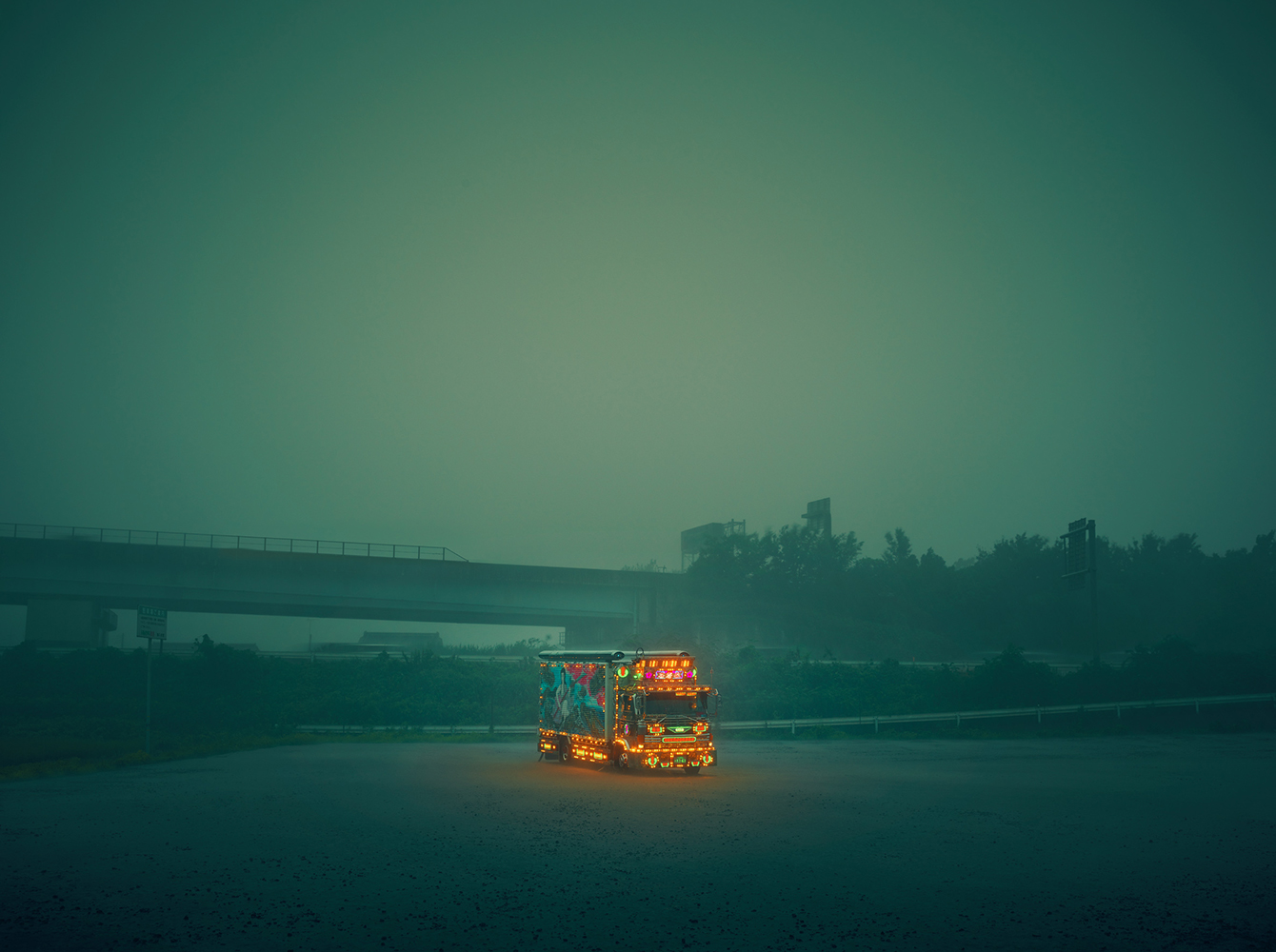 Illuminated truck stands beneath an overpass in a foggy, desolate landscape, bathed in a greenish hue, creating a surreal and atmospheric scene.