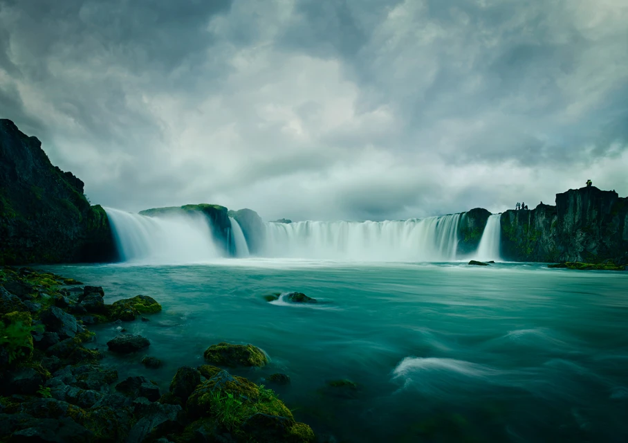 Scenic view of Godafoss waterfall in Iceland with powerful cascades, teal water, and dramatic sky. Lush greenery accents the landscape.