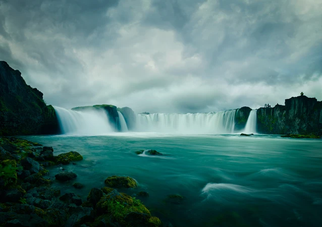 Scenic view of Godafoss waterfall in Iceland with powerful cascades, teal water, and dramatic sky. Lush greenery accents the landscape.