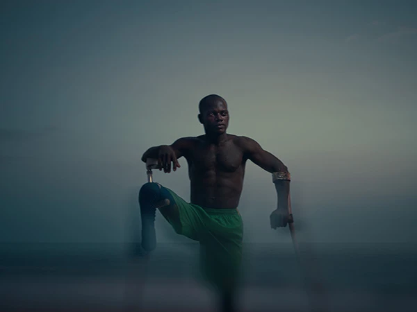 Black male athlete wearing green shorts posing outdoors with crutches against a soft twilight sky and ocean background