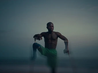 Black male athlete wearing green shorts posing outdoors with crutches against a soft twilight sky and ocean background